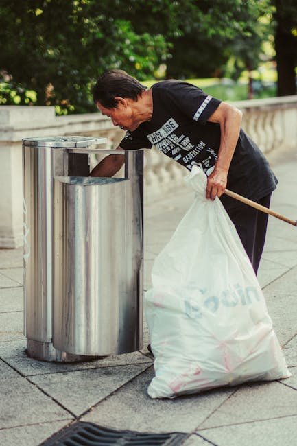 A man with short dark hair, dressed in a black T-shirt and dark pants, is seen disposing of rubbish into a stainless steel litter bin on a paved sidewalk. He is bending slightly forward, with his right hand reaching into the open top of the bin to place or retrieve waste. In his left hand, he holds a large white plastic bag filled with collected rubbish, which appears to be partly crumpled and contains various waste materials. The setting is outdoors, with the background featuring lush green trees and shrubs, and a light-colored stone or concrete wall running parallel to the sidewalk. The environment is well-lit, suggesting daylight, and the scene is indicative of private or independent waste collection, possibly as part of rubbish removal services such as those provided by Rubbish Removal Edgware, aiming to support alternative waste handling outside municipal collection systems.