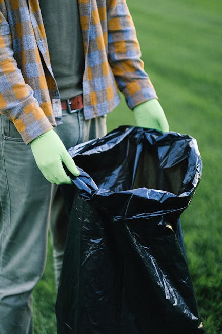 A waste collection operative wearing a high-visibility yellow and orange vest is inspecting the rear of a large red rubbish truck, which is parked at the side of a street. The truck's rear compartment is open, revealing mechanical components and an empty loading area. The worker is standing on the pavement, next to a small waist-high metal barrier with a black garbage bag placed on top. Nearby, there is a motorcycle parked parallel to the truck, partially obstructed by the barrier. The street environment includes a grassy verge with a blue container lying on the ground, and trees and utility poles visible in the background under an overcast sky. The scene suggests an independent waste collection or on-site rubbish removal service, likely part of a local or private waste management effort. The overall atmosphere is neutral and professional, focusing on waste collection activity consistent with rubbish removal services in the UK.