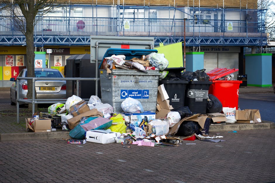 A collection of overflowing rubbish bins and scattered waste materials situated on a paved sidewalk in front of a commercial building. The waste includes black, red, and grey bins filled with mixed household and office rubbish, with some lids open and debris spilling onto the ground. Cardboard boxes, paper, plastic bottles, packaging, and various discarded items are strewn around the bins, forming a cluttered mess. A small car is parked nearby, partially screened by a metal railing, with a license plate visible. In the background, the building features storefronts with closed shutters, and a blue scaffolding structure is visible on the upper level, suggesting ongoing maintenance or construction. The environment appears to be an urban commercial area, with natural daylight illuminating the scene, highlighting the untidy state of the waste collection point. This image illustrates the need for effective rubbish removal services such as those provided by Rubbish Removal Edgware for private or commercial waste management solutions, especially in busy public spaces.