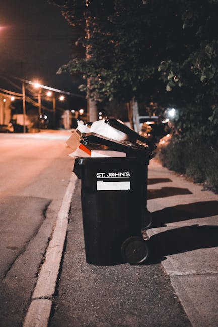 A collection of overflowing rubbish bins and scattered waste materials situated on a paved sidewalk in front of a commercial building. The waste includes black, red, and grey bins filled with mixed household and office rubbish, with some lids open and debris spilling onto the ground. Cardboard boxes, paper, plastic bottles, packaging, and various discarded items are strewn around the bins, forming a cluttered mess. A small car is parked nearby, partially screened by a metal railing, with a license plate visible. In the background, the building features storefronts with closed shutters, and a blue scaffolding structure is visible on the upper level, suggesting ongoing maintenance or construction. The environment appears to be an urban commercial area, with natural daylight illuminating the scene, highlighting the untidy state of the waste collection point. This image illustrates the need for effective rubbish removal services such as those provided by Rubbish Removal Edgware for private or commercial waste management solutions, especially in busy public spaces.
