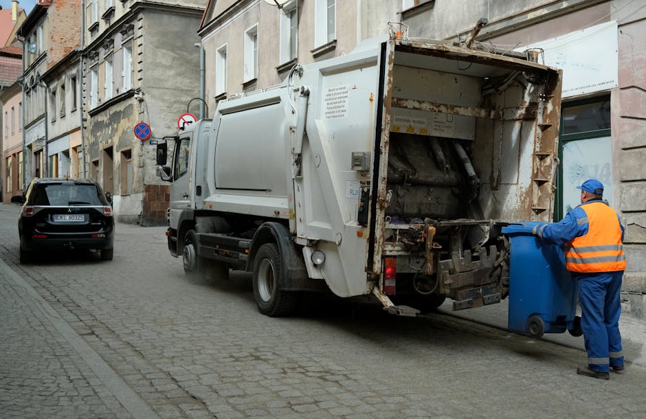 A municipal waste collection vehicle, painted in white with rust and dirt marks on its rear loading compartment, is parked on a narrow cobblestone street in an urban area. The truck's rear open hopper reveals internal mechanical components used for compacting and lifting rubbish. To the right of the vehicle, a sanitation worker dressed in a blue uniform and an orange high-visibility vest is emptying a small blue wheelie bin into the truck's hopper. The worker is wearing gloves and a blue cap, and stands on the cobblestone pavement adjacent to a row of old multi-storey buildings with varied masonry and paint finishes. A black car is parked further along the street on the left side of the image, and there are no other vehicles or pedestrians visible. The scene is lit with natural daylight, capturing the routine process of private waste disposal as part of an alternative rubbish removal service in the area, handled by Rubbish Removal Edgware, demonstrating on-site collection and waste management outside local authority premises.