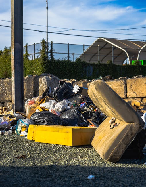 A pile of unmanaged waste positioned on an outdoor gravel surface, consisting of black and white plastic rubbish bags, discarded cardboard and paper, and a yellow plastic tray. A worn and dirty car tire is leaning against the pile, with some loose debris scattered around. Behind the waste, a low stone wall constructed from large, rough-hewn stones separates the area from a background featuring a tall metal pole, overhead power lines, trim green bushes, and a partially visible outdoor structure with a curved roof, likely a sports or storage facility. The scene is illuminated by natural daylight, with a partly cloudy sky overhead, highlighting the informal, unmanaged nature of this rubbish accumulation, which relates to independent waste disposal or rubbish removal services such as those offered by Rubbish Removal Edgware in the context of alternative waste handling outside municipal collection.