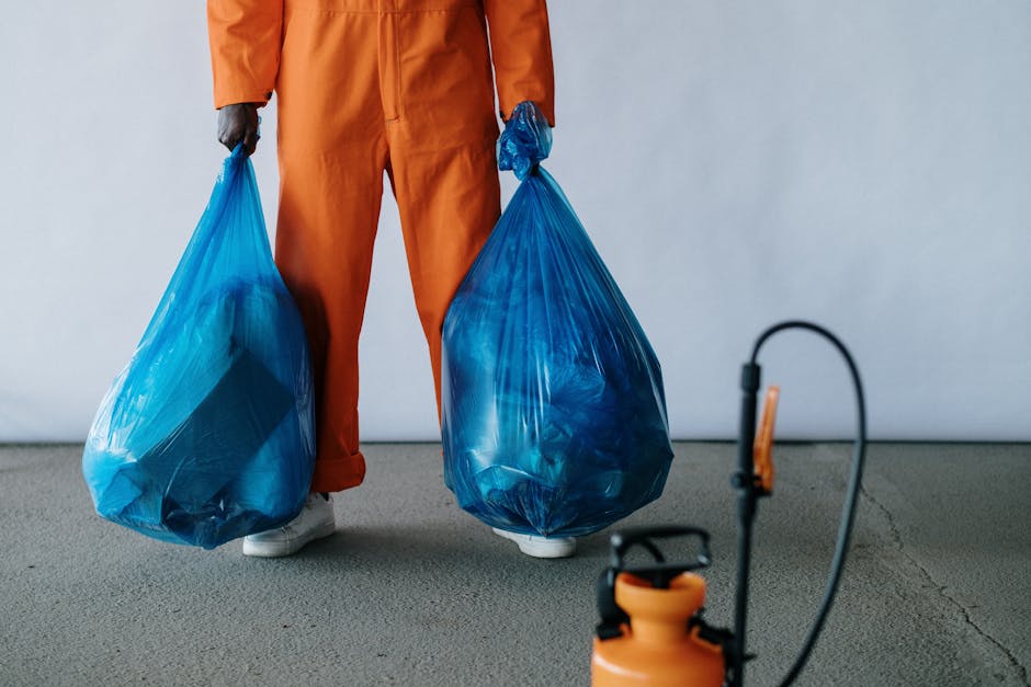 A person wearing an orange protective jumpsuit and white footwear is standing on a plain, grey concrete floor, holding two large, semi-transparent blue plastic rubbish bags filled with waste material. The bags are bulging with discarded items, and their surface reflects light, indicating a sturdy plastic material. The person's hands are gloved, and the background features a smooth, light grey wall, with no other objects or clutter visible. In the foreground, there is a small orange and black sprayer device with a hose attached, placed on the floor. The scene appears to be indoors, possibly in a warehouse or a dedicated waste collection area, with natural and neutral lighting highlighting the bags and the individual’s attire. This setup visually relates to waste collection or rubbish removal services, such as those offered by Rubbish Removal Edgware, focusing on the handling of refuse in an organized and professional manner, suitable for context of private waste disposal or alternative rubbish collection methods outside of municipal services.