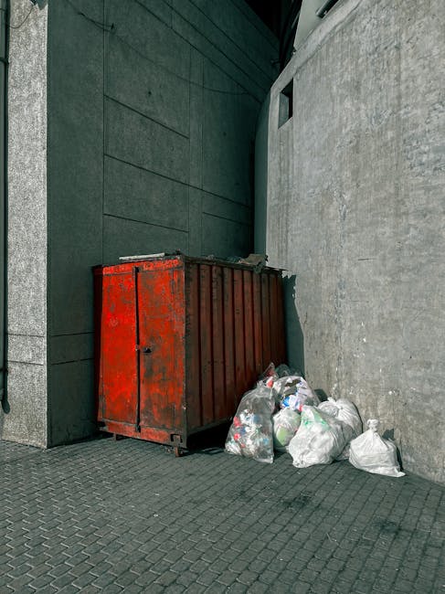 A weathered red metal skip with visible rust and peeling paint is positioned against a grey concrete wall and a dark green building exterior. The skip's surface shows signs of age and exposure to the elements. Several clear plastic bags filled with household waste, including visible plastic bottles and packaging, are piled closely beside the skip on a paved surface. The surrounding environment appears to be an urban side alley or back street area, with the skip placed on a textured asphalt pavement that features a pattern of small, raised diamond shapes. The background consists of the textured concrete wall on the right and a dark facade with horizontal paneling on the left, with a small, narrow window or vent opening seen high on the wall behind the skip. The scene is lit by natural daylight, providing even lighting that highlights the rough textures of the materials, and reflecting the typical setting for private waste disposal or rubbish removal services conducted by companies such as Rubbish Removal Edgware in the context of alternative waste handling options away from public waste collection.